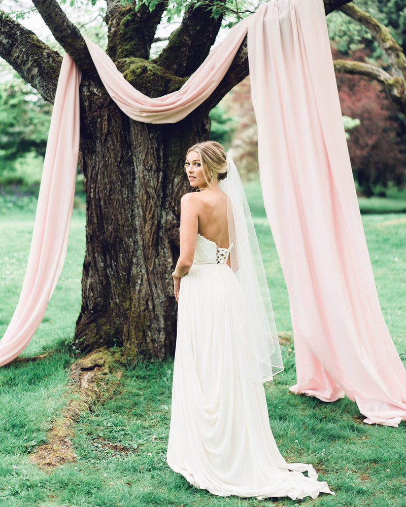 A bride stands in front of a tree draped in blush fabric. She wears a fingertip veil with a thin ribbon trim.