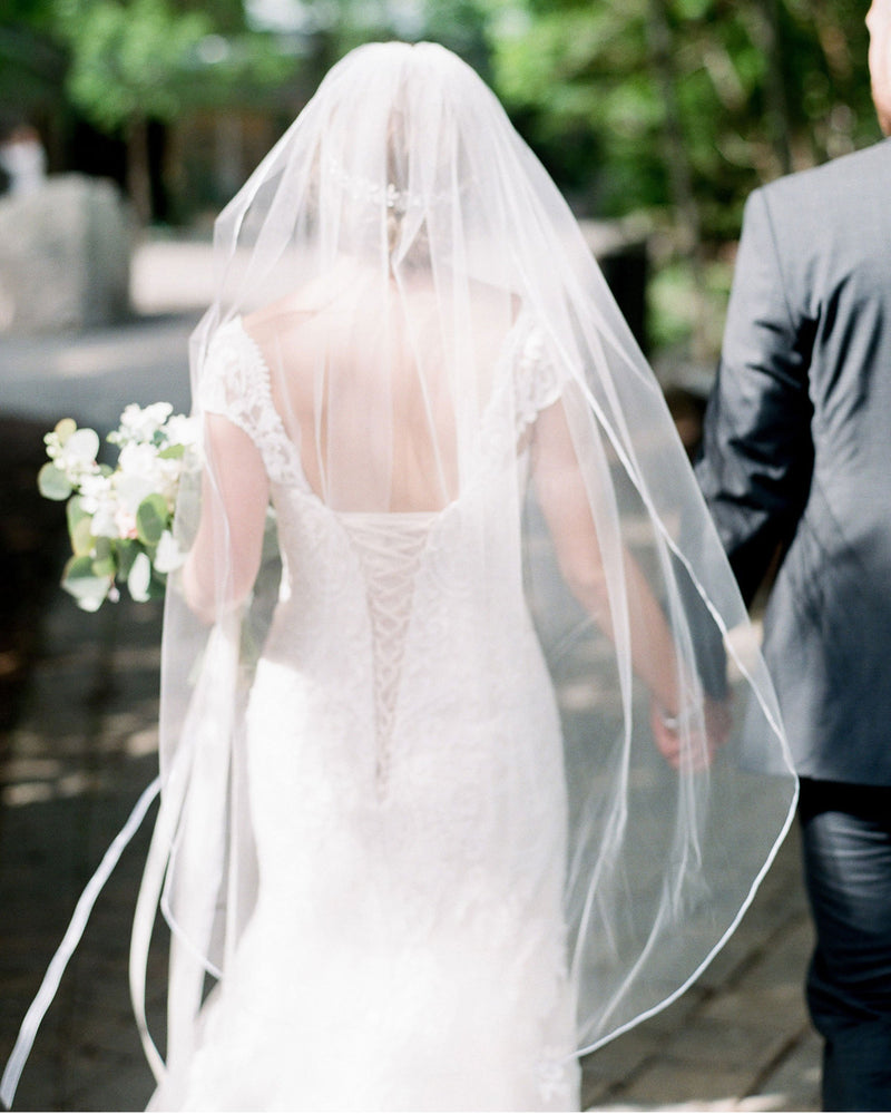 A bride walks away with her groom, wearing the Leila Veil; a fingertip length veil with delicate ribbon trim.