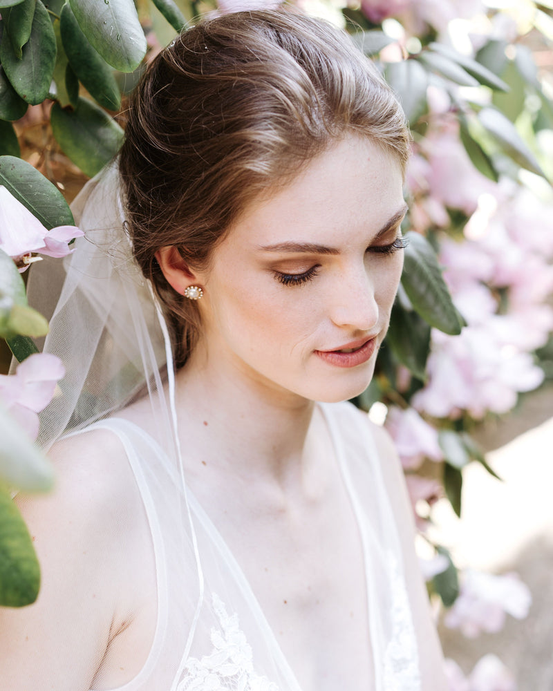 A model is surrounded by flowering trees. She wears the Halo Pearl Stud Earrings with the Leila Ribbon Veil.