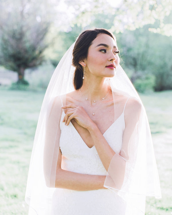 A bride wears a modern pearl jewellery set with Long Pearl Teardrop Earrings and a Layered Pearl Drop Necklace, paired with a two-layer veil with a ribbon trim.